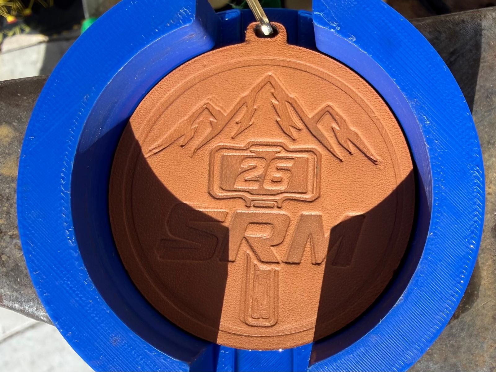 La medaglia della Skyrace del Maglio 2026 con l'emblema SRM e il numero 26 impressi dal colpo di maglio